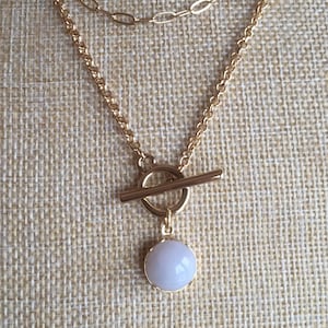 Peut inclure: Un collier doré à plusieurs rangs avec un pendentif rond bleu clair. Le collier comporte deux chaînes, l'une avec un design à maillons délicats et l'autre avec une chaîne classique. Le pendentif est suspendu à un fermoir à bascule.