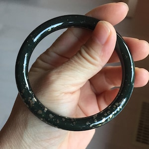 May include: A circular bangle bracelet with a smooth, polished dark green surface. The material features scattered, irregular metallic gold-toned flecks throughout the entire circumference, creating a speckled appearance against the deep green base.