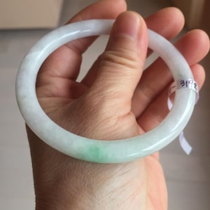 May include: A jade bangle bracelet with a smooth, polished surface. The bracelet is primarily white with a subtle green hue in one area. A small tag with the number "3192" is attached.