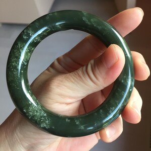 May include: A circular bangle bracelet crafted from dark forest green stone with a polished, smooth surface. The material features natural, lighter green mottled inclusions and cloudy white mineral patterns distributed throughout the solid, thick band.