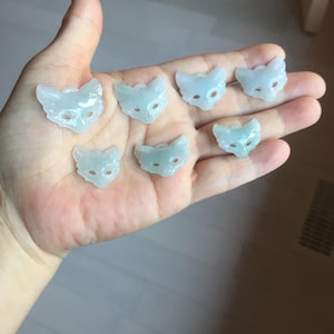 May include: A collection of seven pale green jade cat face pendants. Each pendant features a carved cat face with eye and nose details. The pendants are held in a hand, showcasing their delicate craftsmanship and translucent quality.