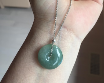 Puede incluir: Collar con colgante redondo de jade verde y cadena de plata. El jade tiene un acabado pulido y brillante con un pequeño agujero en el centro. El collar se muestra sobre un tono de piel claro.