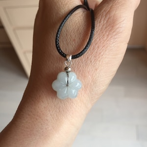 Può includere: Una collana con pendente a forma di fiore azzurro. Il pendente è sospeso a un cordino nero. Il fiore ha cinque petali e un'ansa in metallo argentato. La collana è tenuta in una mano.