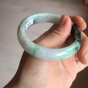 May include: A jade bangle bracelet with a smooth, polished surface. The bracelet features a mix of white and green hues, with the green areas appearing translucent. The bracelet is held in a hand, showcasing its circular shape and natural variations.