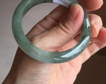 May include: A jade bangle bracelet with a translucent, pale green hue. The smooth, polished surface reflects light, showcasing the natural variations in color. A small white sticker with text is visible on the bracelet.