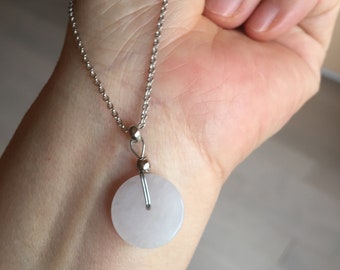 May include: A silver chain necklace with a circular, pale pink pendant. The pendant is smooth and flat, with a small hole in the center. The necklace is displayed on a person's wrist.