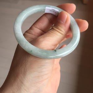 May include: A pale green jade bangle. The smooth, circular bracelet has a translucent appearance with subtle colour variations. A small white tag with text is attached. The bracelet is held in a hand.