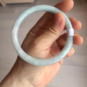 May include: A pale green jade bangle bracelet. The smooth, round bracelet is held in a hand, showcasing its polished surface and subtle color variations. The jade has a translucent quality, with a mix of light green and white hues.