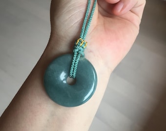 Puede incluir: Un colgante de jade de color verde azulado, con forma circular y un agujero en el centro. Cuelga de un cordón verde azulado con un detalle dorado. El colgante se muestra sobre un brazo.