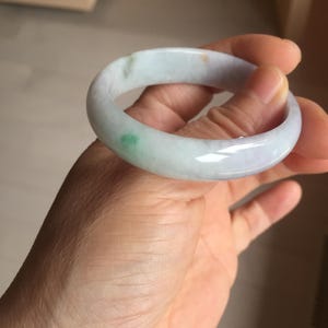 May include: A smooth, rounded bangle bracelet crafted from translucent pale lavender stone. The circular band features subtle, natural inclusions of vibrant green and faint orange hues distributed across its polished, glossy surface.
