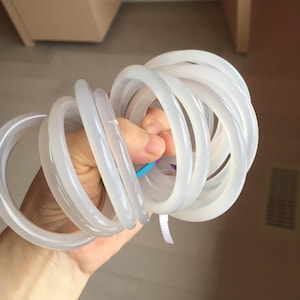 May include: A collection of translucent, white, circular bangles. The bangles are held together, creating a coiled arrangement. The smooth, rounded design suggests a minimalist aesthetic. The light color and shape suggest a jewelry theme.
