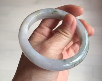 May include: A jade bangle bracelet in shades of white, gray, and green is held in a hand. The smooth, polished surface of the bracelet reflects light, showcasing its natural variations. The circular design is a classic jewelry style.