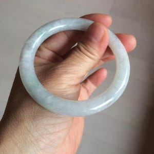 Könnte beinhalten: Ein hellgrünes Jade-Armreif. Das polierte Armband wird in einer Hand gehalten, wodurch sein durchscheinendes Aussehen hervorgehoben wird. Der Jade weist natürliche Farbvariationen auf, mit Bereichen von hellerem und dunklerem Grün.