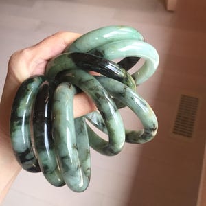 Peut inclure: Plusieurs bracelets en jade de différentes nuances de vert et de noir. Les bracelets lisses et polis sont maintenus ensemble, mettant en valeur leurs motifs naturels et leur finition brillante. Les bracelets sont un bijou classique.