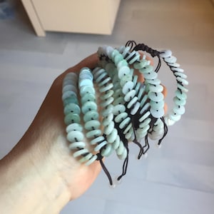 May include: Several jade bracelets with a light green and white color scheme. Each bracelet is made of small, round jade pieces strung together with black cord. The bracelets are stacked together, showcasing their design and color variations.