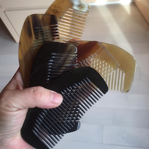 Peut inclure: Trois peignes, dont deux en écaille brune et un en noir. Les peignes sont tenus dans une main.