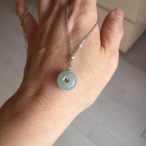 May include: A silver necklace featuring a pale green jade donut pendant. The pendant is round with a hole in the center, suspended from a delicate chain. The necklace is displayed against a neutral background.