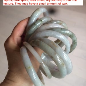 Peut inclure: Plusieurs bracelets en jade jadeite de différentes tailles, tenus dans une main. Les bracelets sont principalement vert pâle avec des inclusions vertes et brunes plus foncées. Le texte de l'image mentionne des défauts.