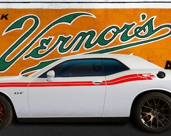 May include: A white Dodge Challenger GT with red and black racing stripes parked in front of a vintage Vernor's Ginger Ale sign. The car has black wheels and tinted windows.