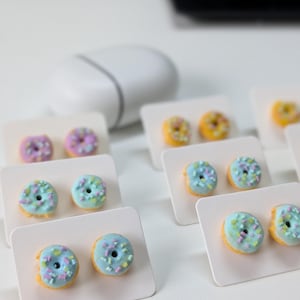 Puede incluir: Un conjunto de pendientes con forma de donut en colores pastel, rosa, azul y amarillo, con chispas. Los pendientes se presentan en tarjetas blancas. En el fondo, un objeto blanco borroso sobre una superficie blanca.