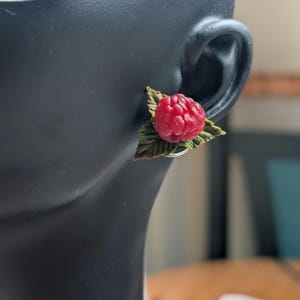 以下が含まれることがあります： 緑の葉が付いた、詳細な赤いラズベリーが特徴のシングルイヤリング。 ラズベリーは質感があり、粘土または同様の素材で作られているようです。 イヤリングはシルバーのポストに取り付けられており、黒いマネキンの頭に着用されています。