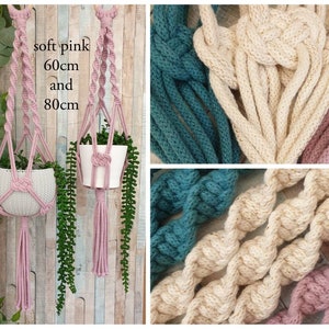 May include: Two pink macrame plant hangers with tassels, one is 60cm long and the other is 80cm long. The hangers are made with soft pink cotton rope and are hanging from a wooden wall. The hangers are holding small potted plants.