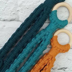 May include: Three macrame plant hangers in teal, dark teal, and orange with wooden rings.