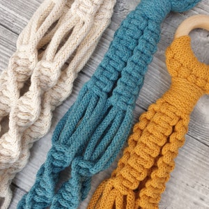 May include: Three macrame plant hangers in different colours: white with gold accents, teal blue, and mustard yellow. Each hanger has a wooden ring at the top.