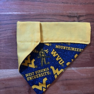 May include: A yellow and blue dog bandana featuring the West Virginia University Mountaineers logo and text. The bandana is folded, showing the yellow top and the blue patterned fabric underneath.