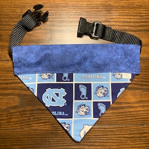 May include: A blue and white patterned dog bandana with the text "CAROLINA" and "UNC" in white. The bandana has a black adjustable strap with two plastic buckles.