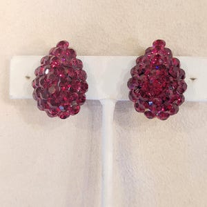 Peut inclure: Une paire de boucles d'oreilles à clip en forme de goutte d'eau, de couleur rouge rubis. Chaque boucle d'oreille est densément recouverte de petits cristaux facettés, créant une surface scintillante et texturée. Les boucles sont présentées sur un support blanc.