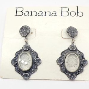 May include: A pair of silver-toned earrings with a floral design. Each earring features a flower-shaped stud, a decorative frame, and a clear oval center with a floral motif. The earrings are displayed on a card with the text "Banana Bob".