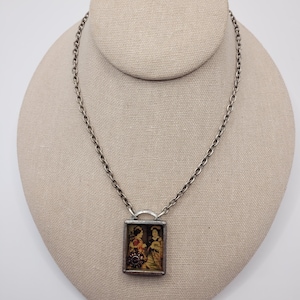May include: A silver-toned metal chain necklace featuring a rectangular pendant. The pendant contains a small, framed image of two figures in traditional attire, accented with a dark, circular decorative element at the bottom left corner.