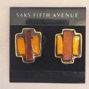 May include: A pair of gold-toned earrings with a geometric design. Each earring features a rectangular shape with amber and brown accents. The earrings are displayed on a black card with the text "SAKS FIFTH AVENUE".