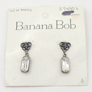 May include: A pair of Banana Bob earrings. Each earring has a decorative top with a dangling, rectangular, clear crystal. The earrings are attached to a white card with the brand name printed in black.