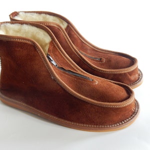 May include: A pair of brown suede slippers with a zipper on the side. The slippers have a soft, fuzzy lining and a brown leather sole.