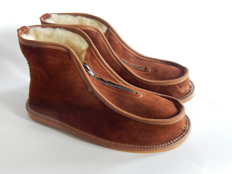 May include: A pair of brown suede slippers with a zipper on the side. The slippers have a soft, fuzzy lining and a brown leather sole.