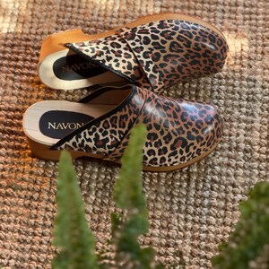 Puede incluir: Un par de zuecos con estampado de leopardo. Los zuecos tienen suela de madera y una parte superior de cuero. El nombre de la marca "NAVON" es visible en la plantilla. Los zuecos son marrones, negros y beiges.