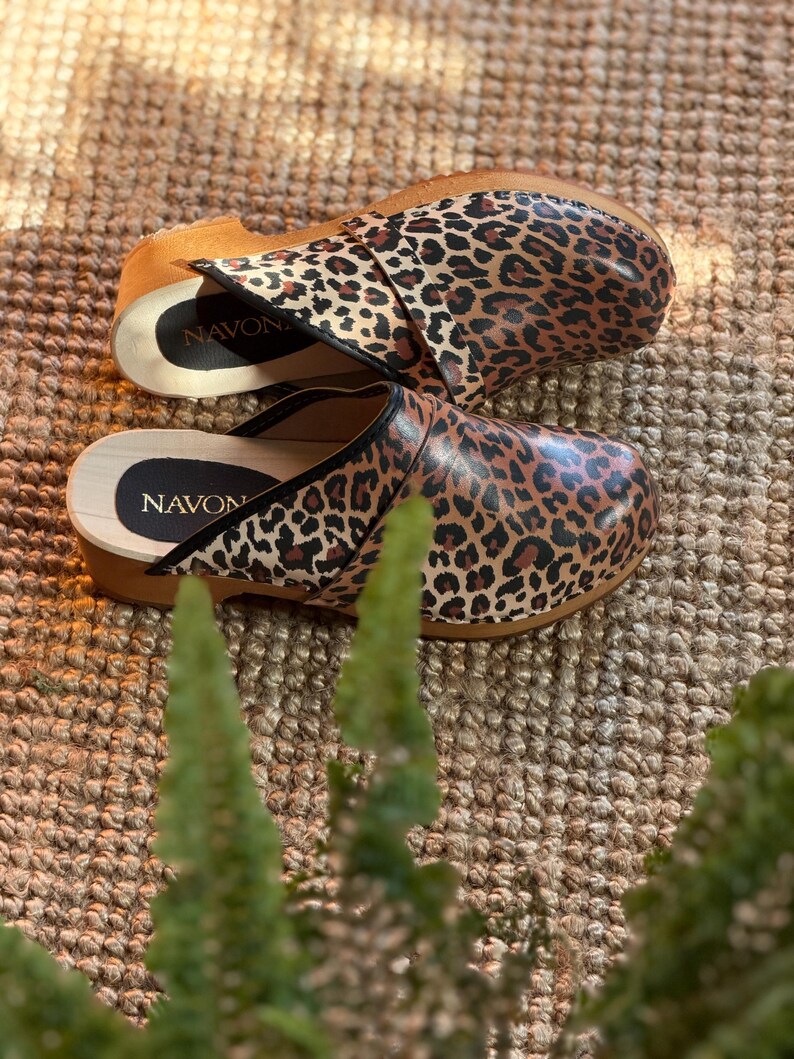 Puede incluir: Un par de zuecos con estampado de leopardo. Los zuecos tienen suela de madera y una parte superior de cuero. El nombre de la marca "NAVON" es visible en la plantilla. Los zuecos son marrones, negros y beiges.