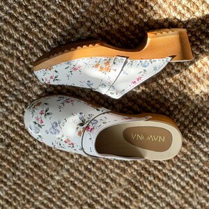 Puede incluir: Dos zuecos blancos con estampados florales, con suela de madera y parte superior de cuero. Los zuecos tienen una base blanca con un delicado estampado de flores rosas, azules y verdes. Las suelas son de madera clara.