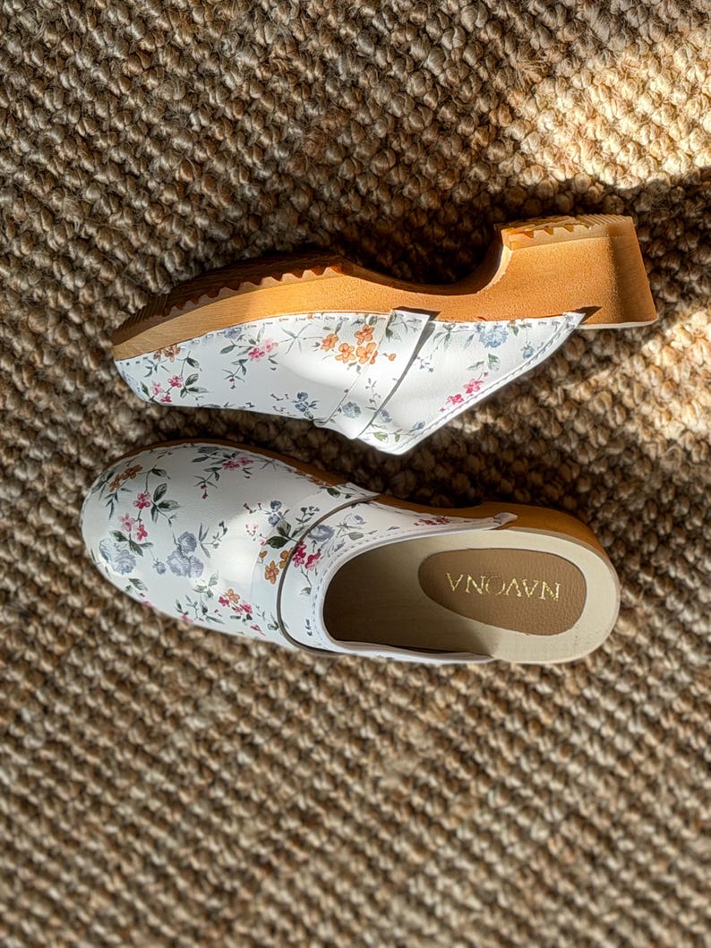 Puede incluir: Dos zuecos blancos con estampados florales, con suela de madera y parte superior de cuero. Los zuecos tienen una base blanca con un delicado estampado de flores rosas, azules y verdes. Las suelas son de madera clara.