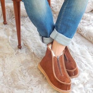 May include: Brown suede slippers with a zipper up the front. The slippers are worn by a person sitting on a chair with their legs crossed. The chair is on a fluffy white rug.