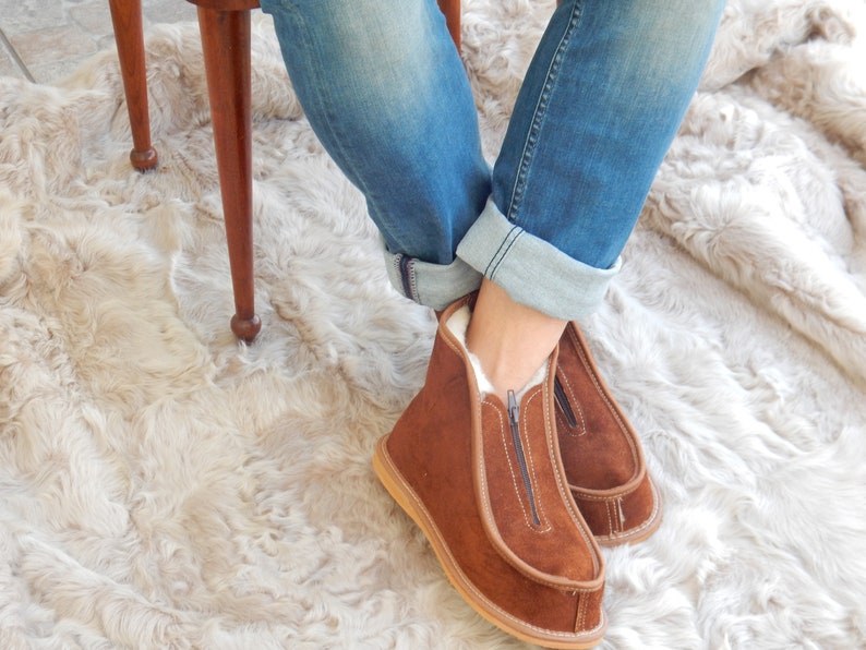 May include: Brown suede slippers with a zipper up the front. The slippers are worn by a person sitting on a chair with their legs crossed. The chair is on a fluffy white rug.