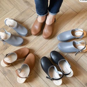 May include: A collection of leather clogs in various colors, including brown, gray, and black, are arranged on a wooden floor. Some clogs have straps, while others are slip-on styles. The clogs are displayed near a person wearing brown clogs.