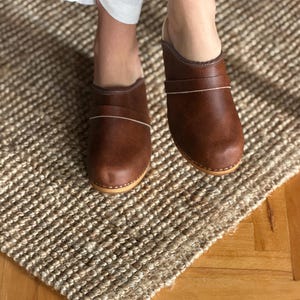 Puede incluir: Zuecos de cuero marr&oacute;n con una l&iacute;nea de acento de color claro. Los zuecos tienen una punta redondeada y una suela de madera. La imagen muestra los zuecos sobre una alfombra tejida.