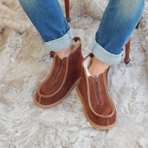 May include: Brown suede zippered boots with a sheepskin lining. The boots are worn by a person sitting on a fluffy white rug.