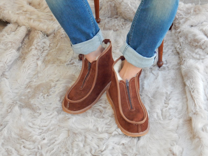 May include: Brown suede zippered boots with a sheepskin lining. The boots are worn by a person sitting on a fluffy white rug.