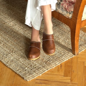 Puede incluir: Zuecos de cuero marrón con suela de color claro y una fina línea blanca. Los zuecos se usan sobre una alfombra de yute tejida, con una silla de madera y una persona que usa pantalones blancos en el fondo.