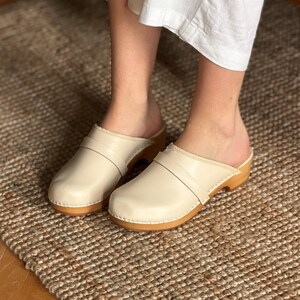 Puede incluir: Zuecos color crema con suela de madera y correa de cuero. Los zuecos se muestran sobre una alfombra tejida. Los zuecos tienen un dise&ntilde;o cl&aacute;sico y minimalista.