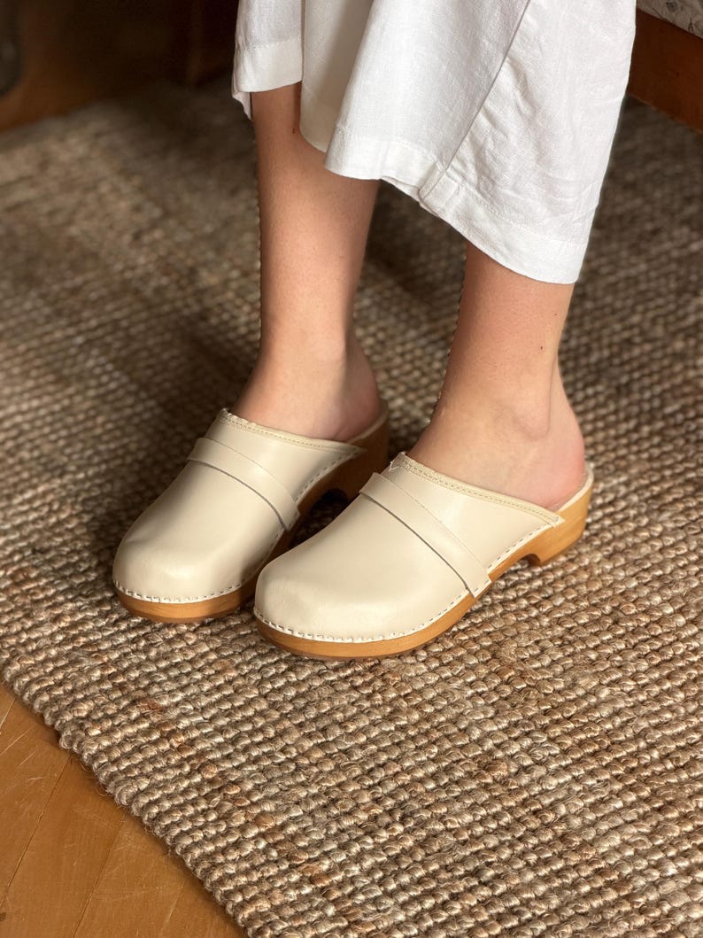 Puede incluir: Zuecos color crema con suela de madera y correa de cuero. Los zuecos se muestran sobre una alfombra tejida. Los zuecos tienen un dise&ntilde;o cl&aacute;sico y minimalista.